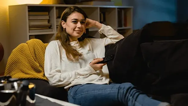 A woman in a white jumper and jeans is relaxing on a sofa with a remote control, watching TV in a cosy living room with shelves behind her.