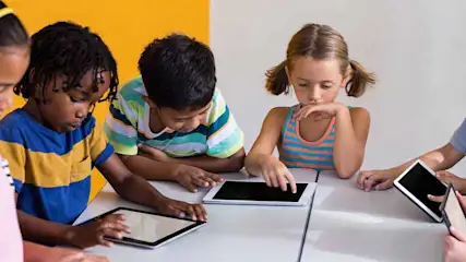 Meerdere kinderen zitten aan een tafel met tablets voor een gele en witte achtergrond.