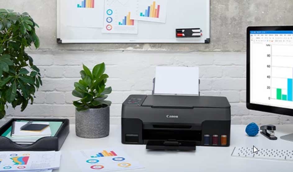 Een zwarte Canon printer staat op een bureau met planten, grafieken en een computerscherm. Het Canon-logo is goed zichtbaar.