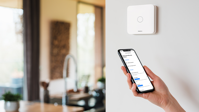 Witte schakelaar aan de muur, hand houdt telefoon met app. Achtergrond: keuken met raam, aanrecht en plant.