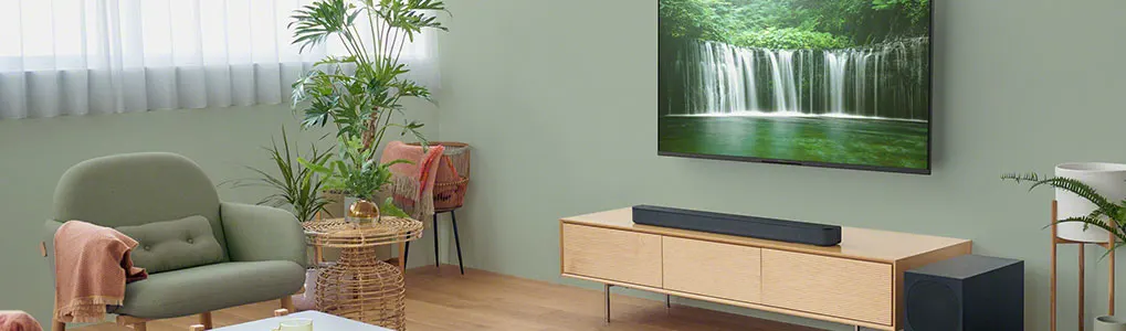 Een sfeervolle zithoek met zachtgroene muur, zwevend houten dressoir met zwarte soundbar en subwoofer, bovenop de kast een tv die een waterval toont, daarnaast een rotan bijzettafeltje, fauteuil en grote planten.