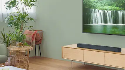 Een sfeervolle zithoek met zachtgroene muur, zwevend houten dressoir met zwarte soundbar en subwoofer, bovenop de kast een tv die een waterval toont, daarnaast een rotan bijzettafeltje, fauteuil en grote planten.