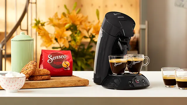 Een zwarte Senseo-koffiemachine met twee kopjes koffie, een pak koffiepads, koekjes en een pot op een aanrecht.