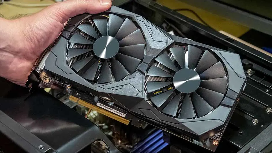 Een hand houdt een grijze grafische kaart vast met twee ventilatoren, boven een open computerkast.