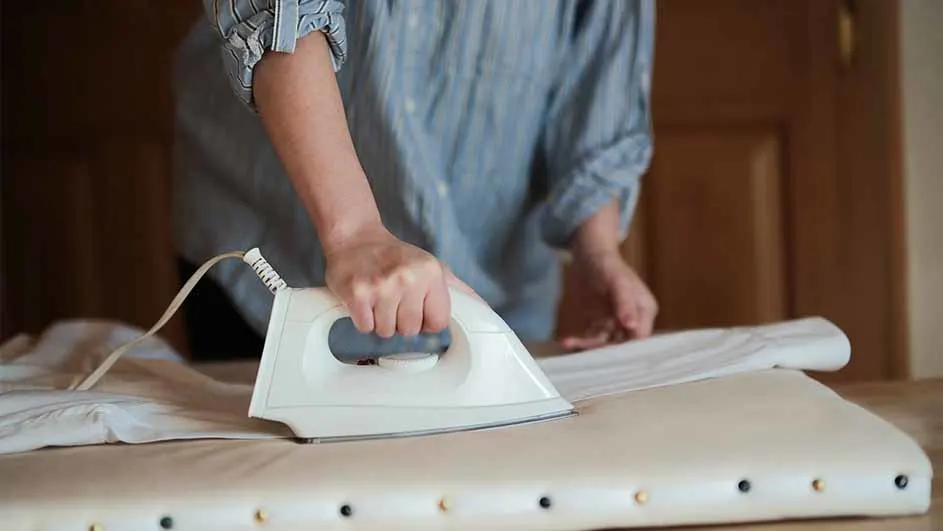 Een persoon strijkt een wit kledingstuk met een wit strijkijzer op een gestreken plank. Achtergrond is een houten deur.