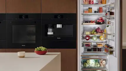 Een moderne keuken met Miele inbouwapparatuur en een goed gevulde koelkast. Boven de koelkast staan voorraadpotten met pasta. De houten kasten geven een warme uitstraling.