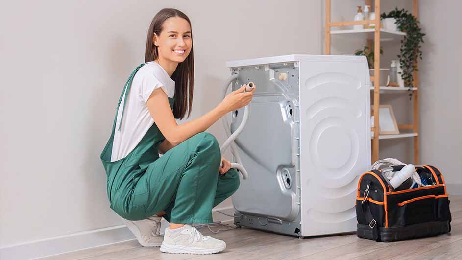 A smiling woman in green overalls kneels by a white washing machine, holding a hose. A toolbox sits nearby on the wood floor.