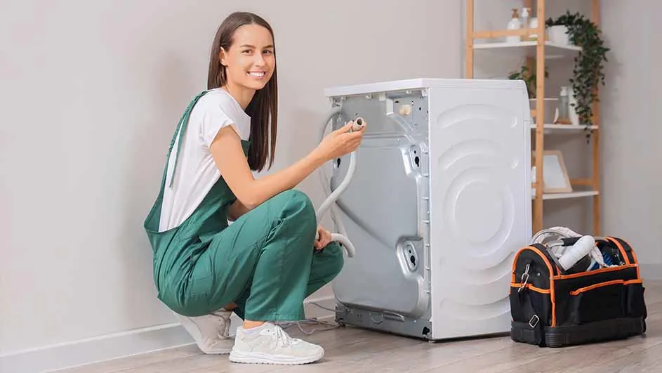 A smiling woman in green overalls kneels by a white washing machine, holding a hose. A toolbox sits nearby on the wood floor.