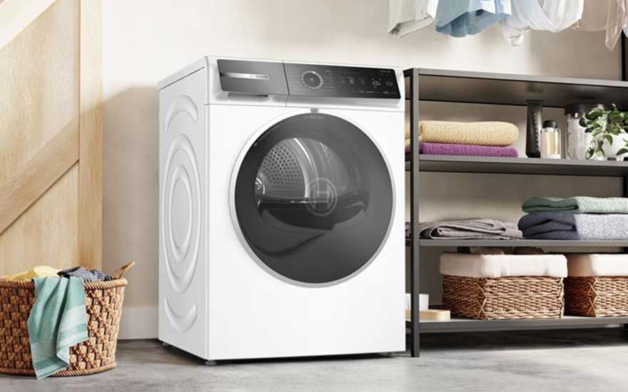 Vrijstaande Bosch wasdroger met een donker venster aan de voorkant, naast rekken vol handdoeken en een wasmand in een lichte ruimte.