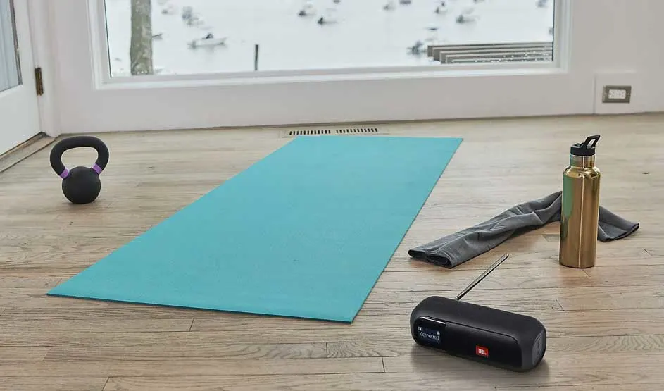 Op een houten vloer ligt een blauwe yogamat met een kettlebell, een handdoek, een gouden drinkfles en een zwarte JBL radio.