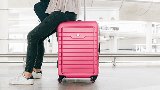 Een persoon zit op een roze koffer in een wachtruimte met een groot raam op de achtergrond.