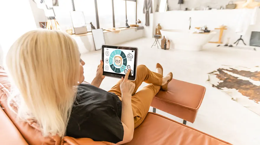 Vrouw op een bruine bank met een tablet, waarop een app te zien is voor huisautomatisering. Op de achtergrond een lichte kamer.