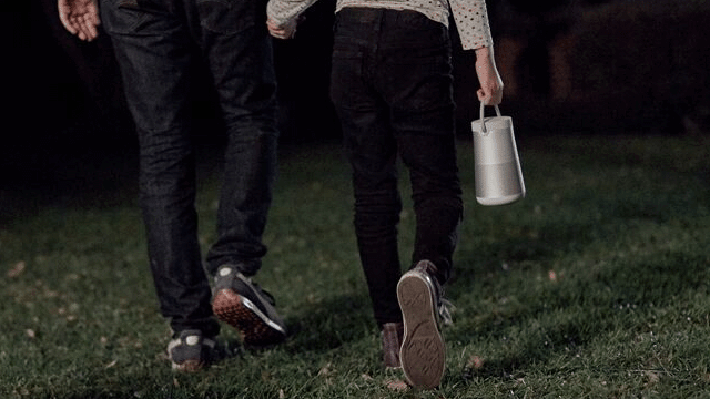 Twee personen lopen over gras. Een persoon draagt een zilverkleurige draagbare speaker. Beide dragen donkere broeken en schoenen.