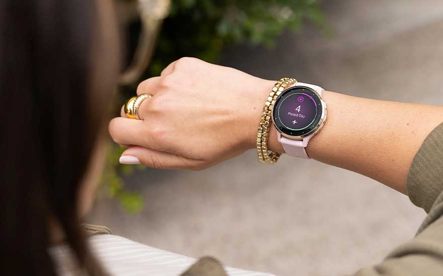Op de pols van een vrouw zie je een moderne smartwatch met roze band naast een gouden armband, het ronde scherm toont een duidelijke melding.