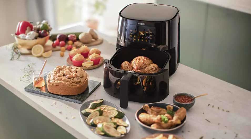 A black air fryer cooks food on a counter with plates of vegetables, fried chicken, cake, and fruit.
