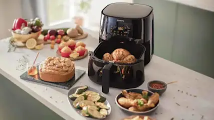 A black air fryer cooks food on a counter with plates of vegetables, fried chicken, cake, and fruit.
