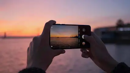 Twee handen houden een telefoon vast die een zonsondergang over water fotografeert. Op de achtergrond vaart een boot.