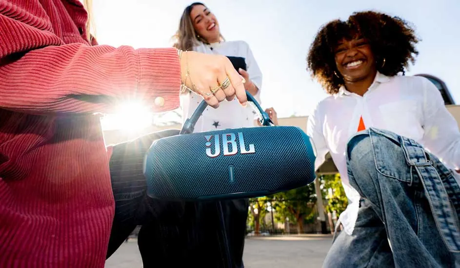 Drie vrienden lachen buiten in de zon terwijl iemand een blauwe JBL draagbare luidspreker omhooghoudt, klaar om samen muziek te luisteren.