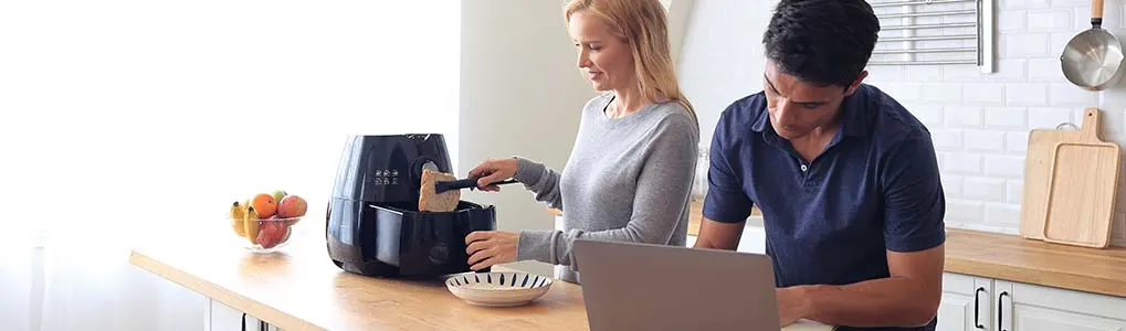 Een persoon bedient een airfryer op een keukenblad, terwijl een andere persoon op een laptop werkt. De keuken heeft een lichte, moderne inrichting met planken, kookgerei en een fruitschaal op het aanrecht.