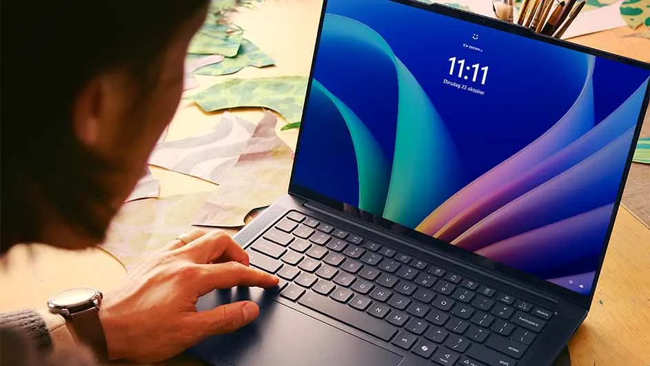 Laptop met blauw scherm waarop 11:11 staat, omringd door een persoon, penselen en papieren vormen.