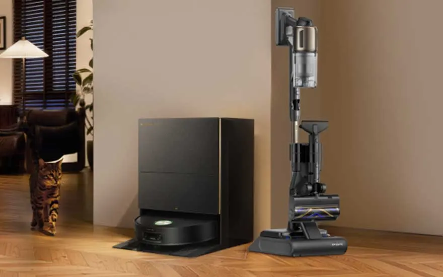 Een robotstofzuiger met dockingstation en een steelstofzuiger staan tegen een beige muur, terwijl een kat binnenloopt.