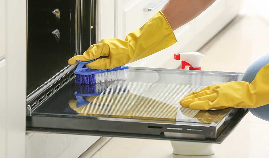 Iemand met gele handschoenen schrobt de glazen ovendeur schoon met een borstel en een doek ernaast.