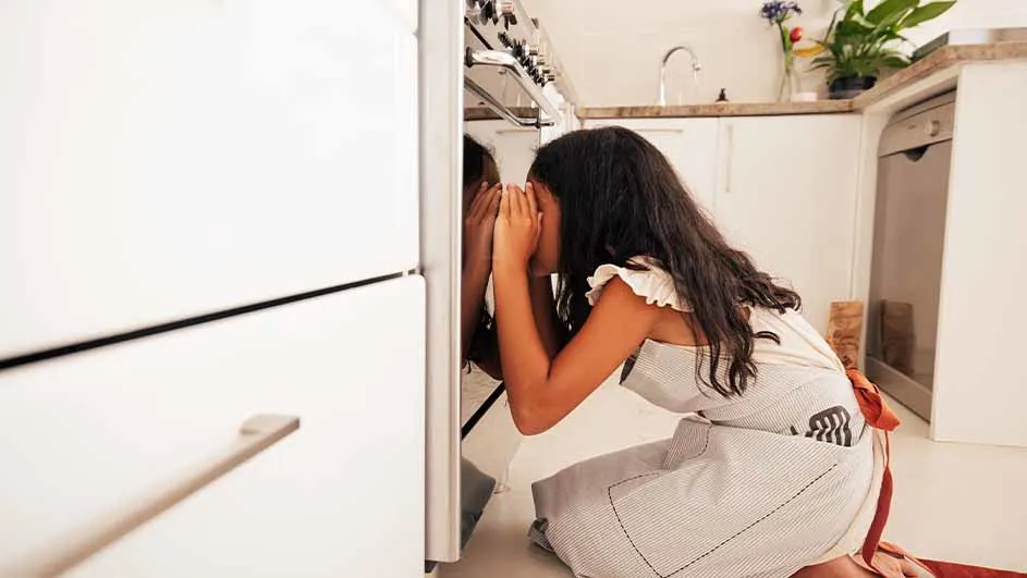 Een vrouw knielt voor een oven in een keuken, kijkend naar binnen met haar handen tegen de ovendeur.