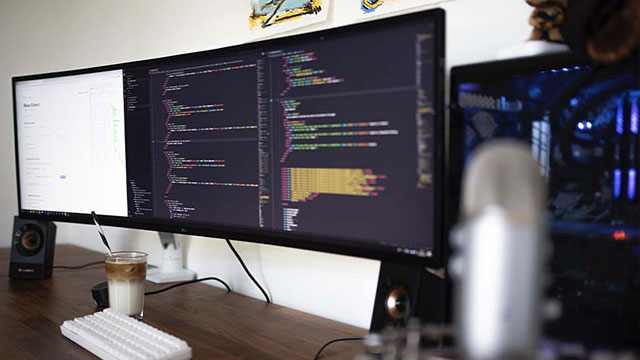 Een bureau met een toetsenbord, een drankje, luidsprekers, een microfoon en een monitor met code.