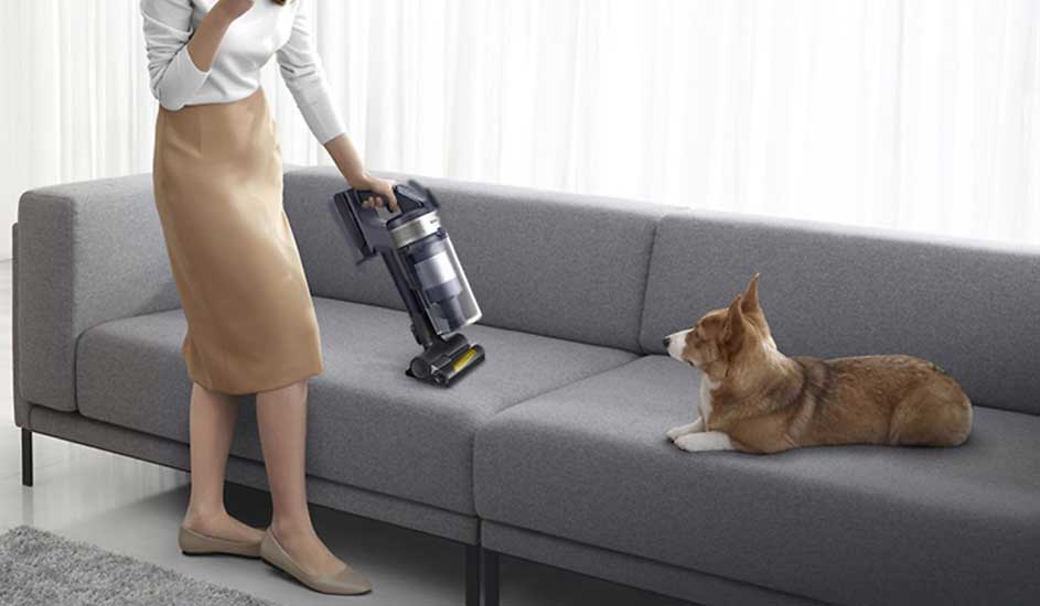 Een vrouw stofzuigt een grijze bank met een Samsung stofzuiger terwijl een hond rustig naast haar ligt. Merk: Samsung.
