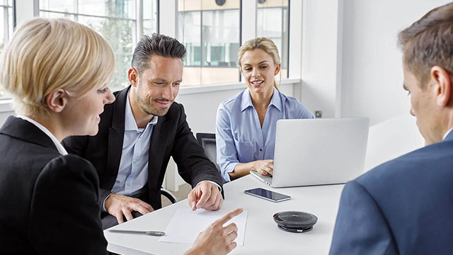 Vier personen in pak aan een witte tafel met een laptop, telefoon, pen en speakerphone.