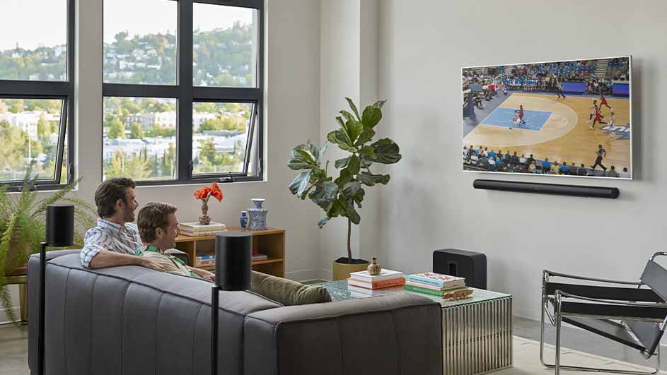 Twee mannen zitten op een grijze bank en kijken naar een basketbalwedstrijd op tv in een woonkamer.