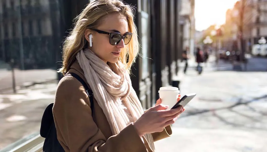 Vrouw met zonnebril en koptelefoon houdt een telefoon en een beker vast op een straat met gebouwen.