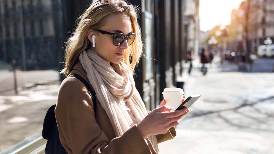 A person in sunglasses and scarf holds a phone and cup outside. Buildings are visible in the background.