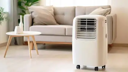 Een witte mobiele airco staat in een kamer met een beige bank en een ronde tafel met decoratieve objecten.