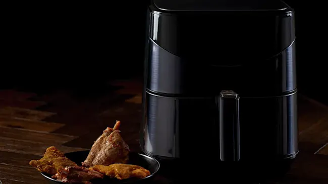 A black air fryer sits near a black bowl containing cooked food on a wooden surface against a dark background.