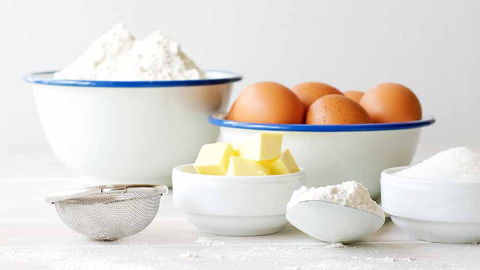 Witte kommen gevuld met bloem, eieren, boter en suiker, plus een metalen zeef, op een witte houten ondergrond.