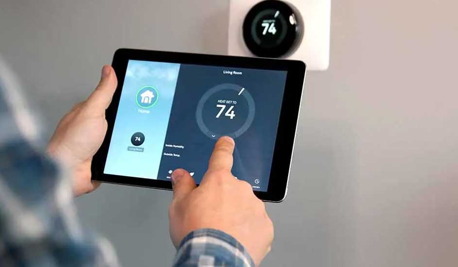 Hand holds a tablet showing a thermostat set to 74 degrees. A round thermostat is mounted on the wall above.