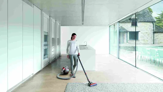Vrouw stofzuigt in een moderne kamer met een stofzuiger. Buiten is een stenen huis te zien door een grote glazen wand.