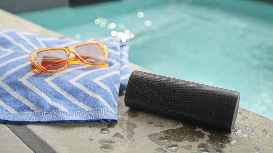 Een zwarte Sonos speaker, een blauw-witte handdoek en een oranje zonnebril liggen naast een zwembad op een betonnen oppervlak.