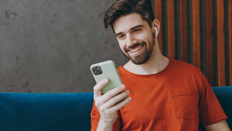 Een man met een baard glimlacht en kijkt naar een groene smartphone. Hij draagt een oranje shirt en oortjes, zit op een blauwe bank.