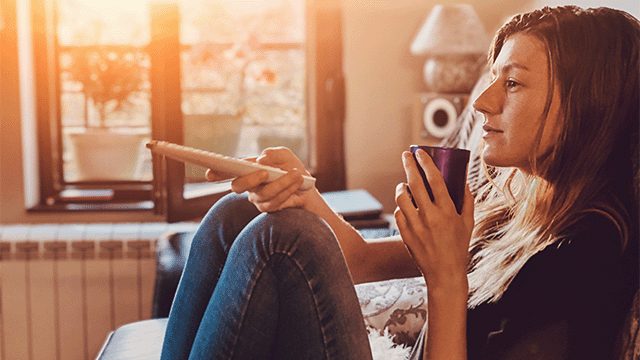 Een vrouw zit op een bank met een kopje en een afstandsbediening in haar handen, bij een raam.