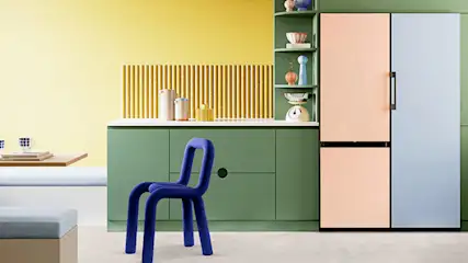 Een keuken met gele muren, groene kasten en een koelkast met roze en blauwe panelen. Een blauwe stoel staat voor het aanrecht.