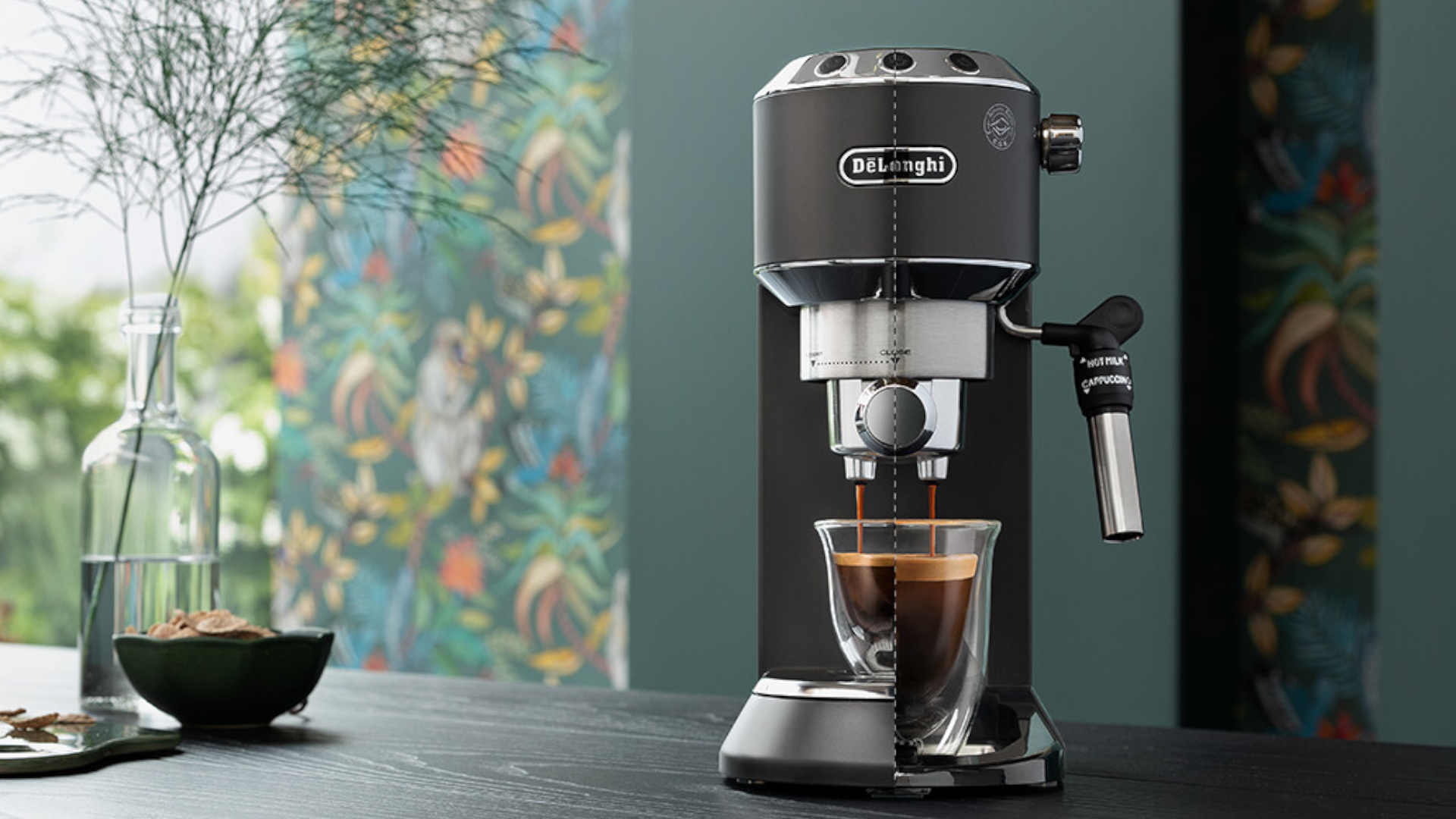 Een DeLonghi espressomachine vult een glazen kop met koffie op een donkerhouten tafel met een kleurrijke achtergrond.