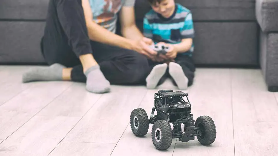 Een vader en kind spelen met een zwarte speelgoedauto op een houten vloer, met een grijze bank op de achtergrond.