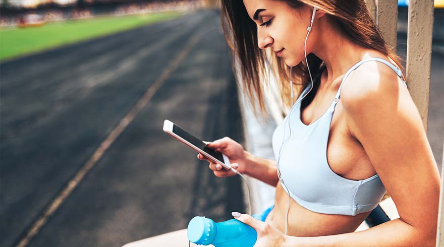 Een vrouw in sportkleding gebruikt een telefoon met oortjes, met een blauwe fles in de hand, op een atletiekbaan.