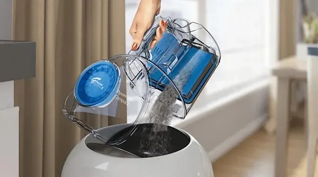 A hand empties a clear vacuum dust canister into a round bin, grey debris pouring out inside a bright, tidy modern room.