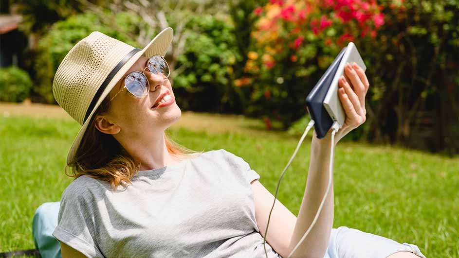 Vrouw met hoed en zonnebril ligt in gras, laadt telefoon op met powerbank. Achtergrond: groene struiken met rode bloemen.