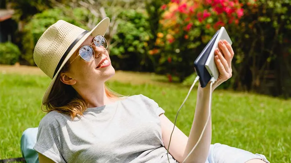 Vrouw met hoed en zonnebril ligt in gras, laadt telefoon op met powerbank. Achtergrond: groene struiken met rode bloemen.
