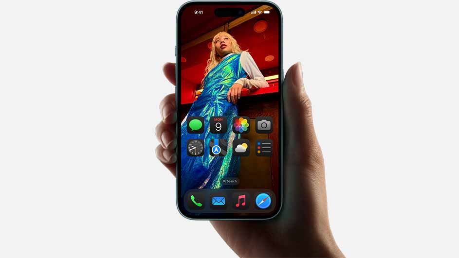Een hand houdt een telefoon vast met een homescreen. Op de achtergrond van het scherm staat een persoon in blauwe kleding.