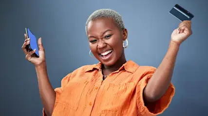 Een lachende persoon met oorbellen, een oranje overhemd, een telefoon en een creditcard tegen een grijze achtergrond.